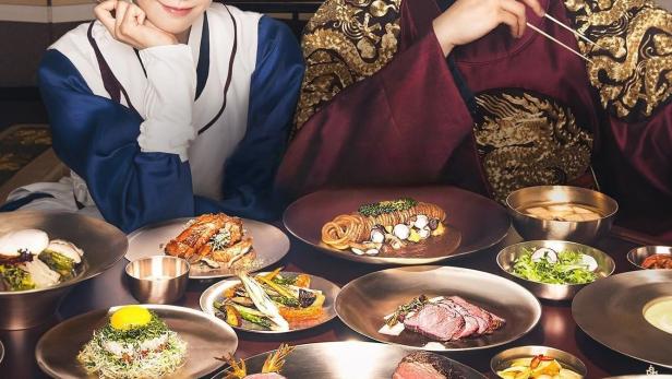 Ein Mann und eine Frau in traditioneller koreanischer Kleidung sitzen an einem Tisch voller Speisen.