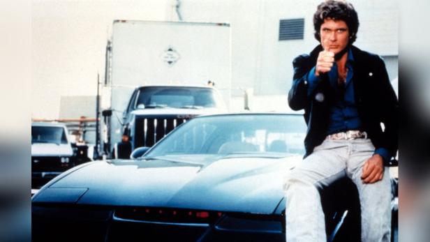 David Hasselhoff sitzt auf der Motorhaube des Auto K.I.T.T. in der Kultserie „Knight Rider“ und streckt den rechten Daumen in die Höhe.