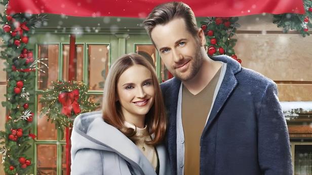 Ein Paar steht vor einem Banner für den Film „Buchladen der Liebe: Winterzauber für zwei Herzen“.