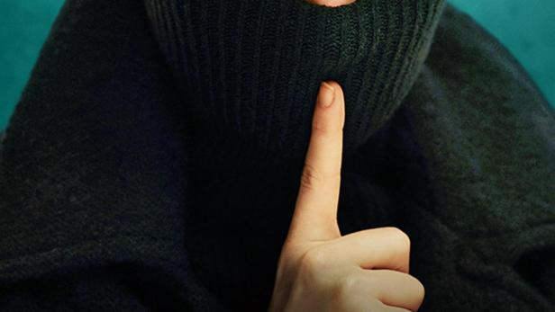 Eine Frau mit rotem Haar hält einen Finger vor den Mund, auf dem Bild steht „Say Nothing“.