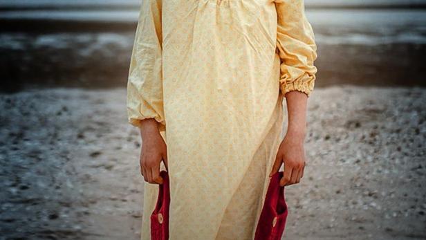 Ein Mädchen im gelben Kleid steht am Strand und hält rote Schuhe in den Händen.