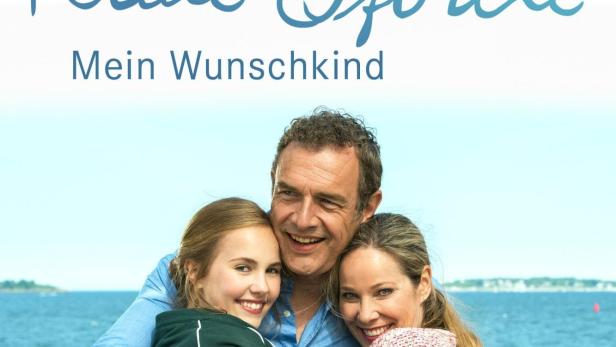 Das Filmplakat zu „Katie Fforde: Mein Wunschkind“ zeigt drei lächelnde Personen vor einer Küstenlandschaft.