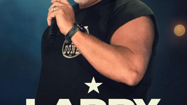 Larry the Cable Guy steht mit einem Mikrofon auf der Bühne.