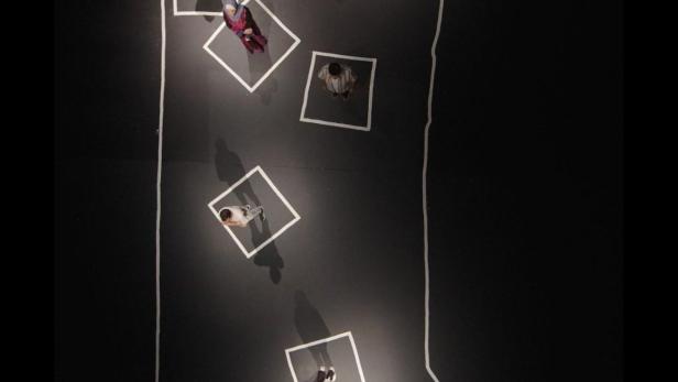 Eine Draufsicht zeigt Menschen, die in weißen Quadraten auf einer schwarzen Fläche stehen, die von einer weißen Linie umrandet ist.