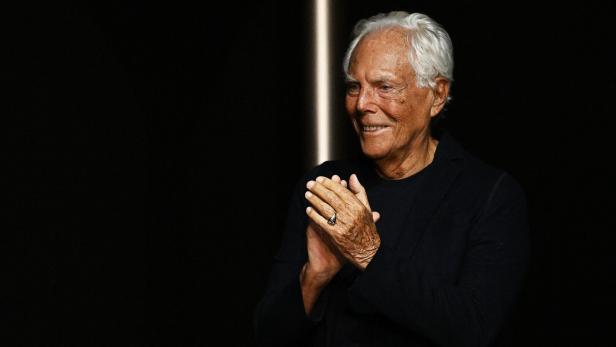 Giorgio Armani lächelt und klatscht vor dunklem Hintergrund Beifall.