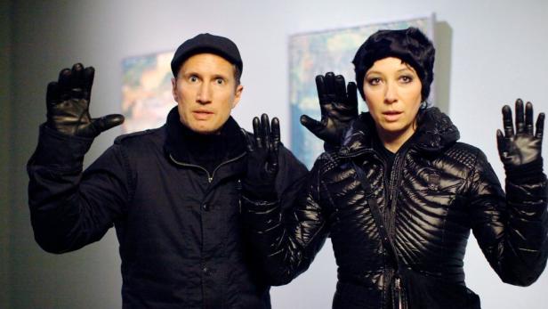 Benno Fürmann und Ursula Strauss in schwarzer Kleidung heben die Hände vor Gemälden an der Wand.