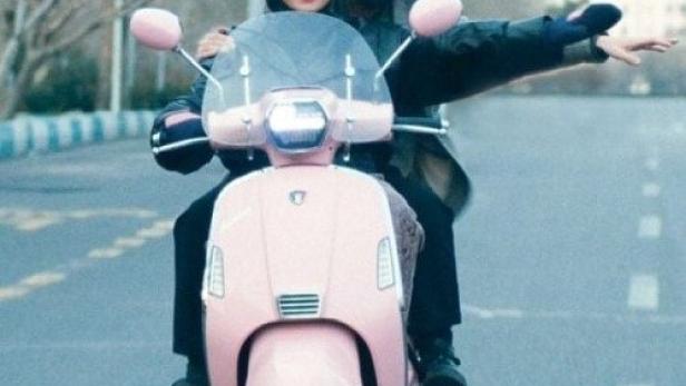 Ein Paar fährt mit einem rosa Motorroller auf einer Straße.