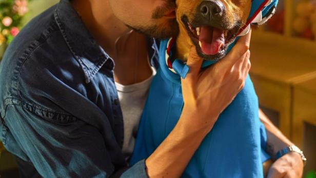 Ein Mann umarmt einen Hund, der ein blaues Haikostüm trägt. Im Vordergrund steht der Titel „Caramelo“.