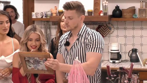 „Sommerhaus der Stars“-Kandidaten stehen in der Küche und betrachten die Rückseite eines gerahmten Bildes.