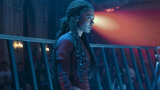 Serienfigur Marie steht hinter einem Geländer in einem mit Scheinwerfer erhelltem Raum.