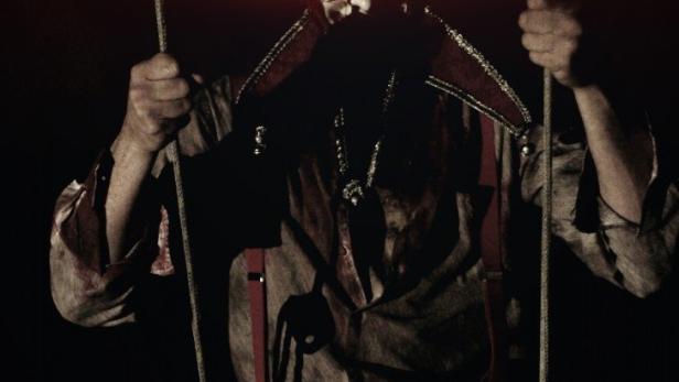 Ein unheimlicher Narr mit leuchtend roten Augen hält Seile in den Händen.