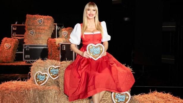 Heidi Klum sitzt im Dirndl mit Lebkuchenherz in Händen auf einem Strohballen.