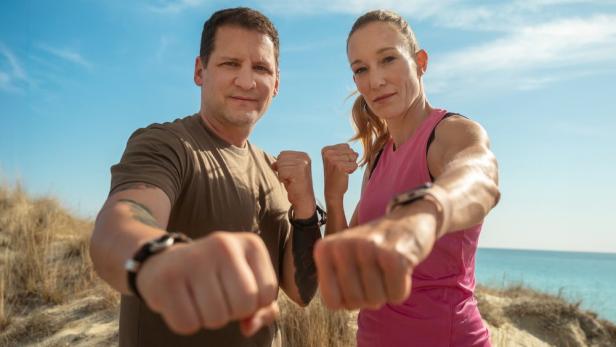 Moderatoren Ramin Abtin und Christine Theiss strecken uns in Sanddünen am Strand ihre Fäuste entgegen.