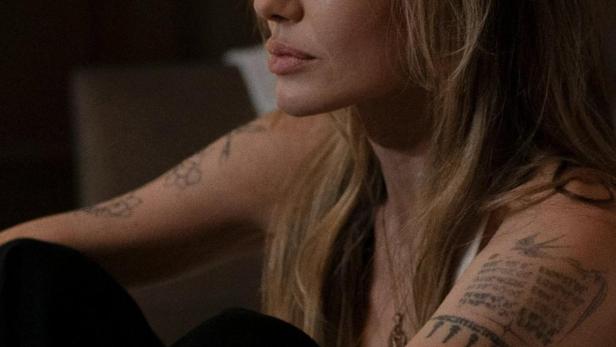 Angelina Jolie sitzt mit Tattoos auf den Armen und blickt zur Seite.