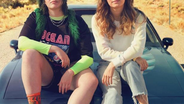 Zwei junge Frauen sitzen auf der Motorhaube eines Autos vor einer Wüstenlandschaft.