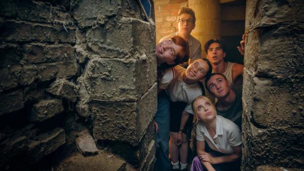 Sechs junge Hauptfiguren sehen in einem Keller durch einen gemauerten Spalt