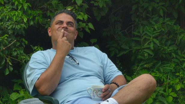 Silva Gonzalez sitzt zurückgelehnt im Grünen mit Kaffeetasse in der Hand und Zeigefinger am Mund