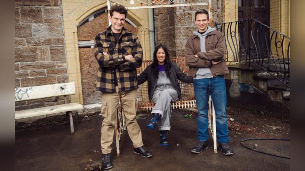 Frau sitzt auf alter Metallbank im Freien, zu ihren beiden Seiten steht je ein Mann