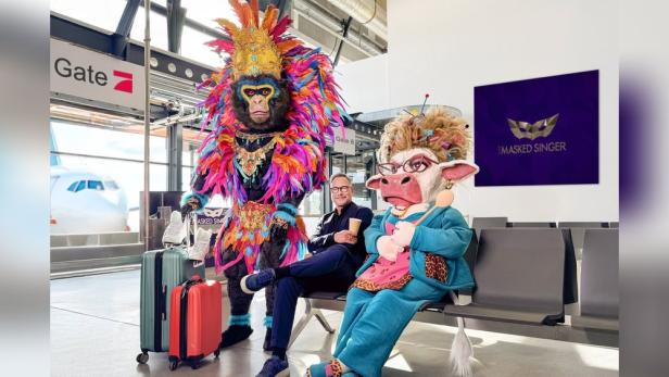 Mann sitzt am Flughafen zwischen zwei als Affe und Kuh Kostümierten