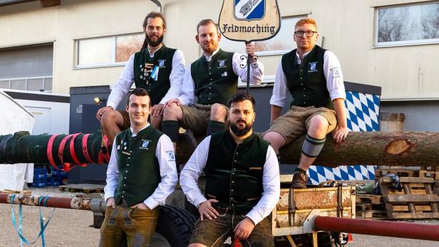 Fünf junge Männer in Lederhosen sitzen auf einem Maibaumstamm auf einem Anhänger