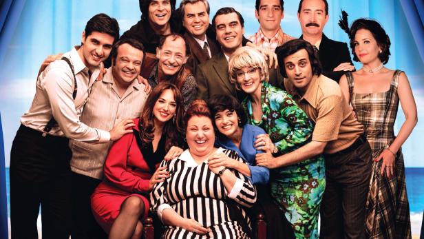 auf einer Theaterbühne ist ein ganzes Ensemble um eine sitzende ältere Frau versammelt