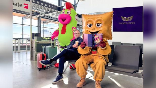 Moderator sitzt am Flughafen zwischen zwei Masked-Singer-Figuren