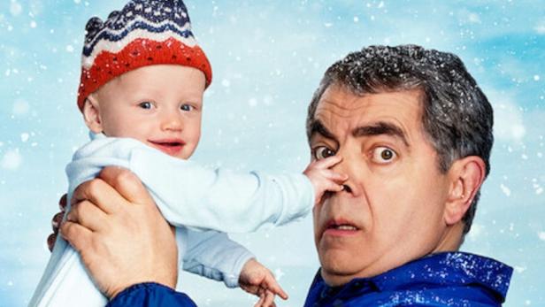 Rowan Atkinson hält vor sich ein Baby in die Höhe, das ihm an die Nase fasst