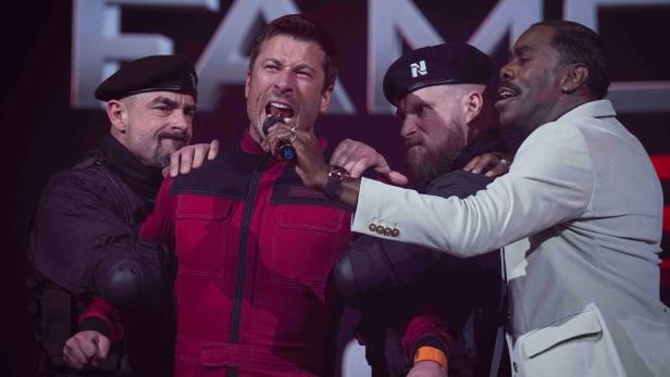 Mann in rotem Dress wird von zwei Uniformierten festgehalten, während er in ein Mikro brüllt