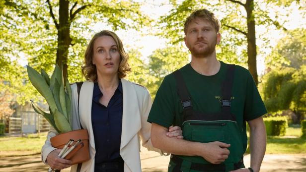 Blinde Frau mit Topfplanze im Arm wird von einem Mann durch den Garten geführt