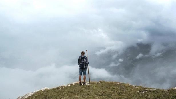 Mann mit Bergstock steht auf grasigem Gipfel und blickt ins fast ganz von Wolken verdeckte Tal