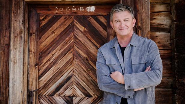 Hans Sigl lehnt als "Der Bergdoktor" an der Holztür einer Hütte