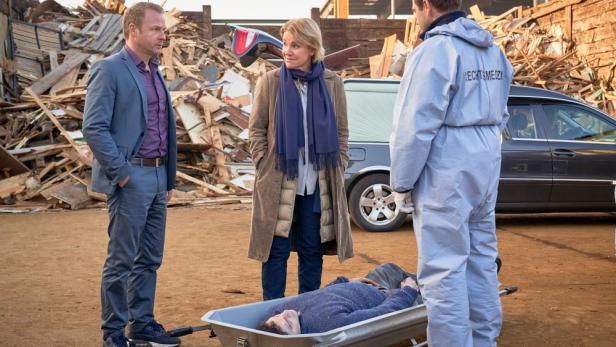 Drei Personen stehen auf einem Recyclinghof um einen Toten Mann, der auf dem Boden in einer Wanne liegt