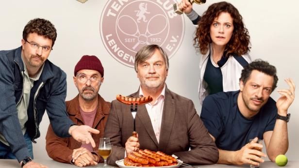 Hape Kerkeling, Christoph Maria Herbst und weitere Darsteller sitzen an einem Tisch mit Teller voller Grillwürsten unter Tennispokalen 