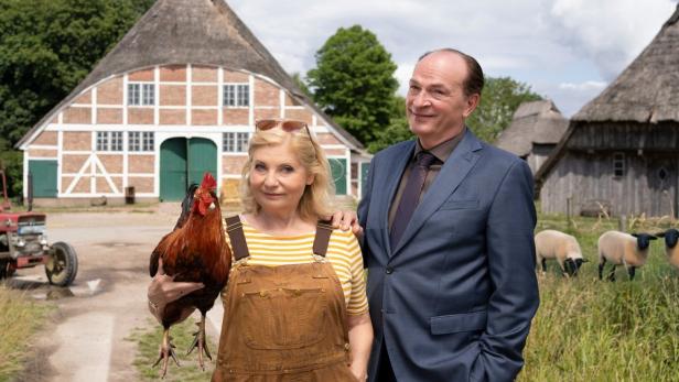 Mann legt vor einem Bauernhof seine Hand auf Schulter einer Frau, die ein Huhn im Arm hält