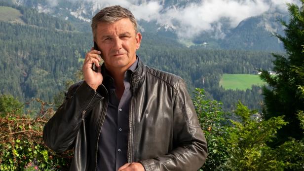 Hans Sigl in seiner Bergdoktor-Rolle mit Handy am Ohr vor Waldkulisse