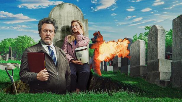 Nicholas Ofczarek steht zur Hälfte in einem ausgeschaufelten Grab, Julia Jentsch sitzt dahinter vor Grabstein mit Urne in der Hand