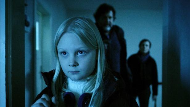 Mädchen geht durch einen düsteren Hausflur, zwei Erwachsene folgen