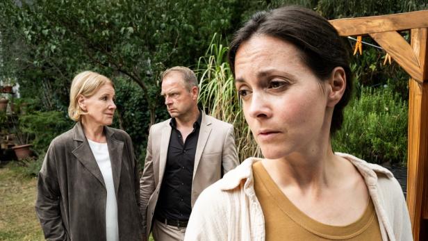 Frau im Vordergrund blickt in Garten geistesabwesend zur Seite, im Hintergrund blicken Mann und Frau einander an