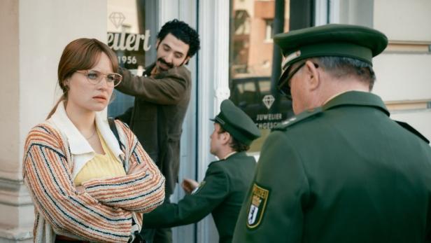 Frau steht mit verschränkten Armen vor einem Polizisten, während im Hintergrund ein anderer eine Verhaftung vornimmt