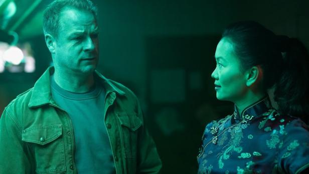 Mann steht in grünlicher Beleuchtung einer asiatischen Frau gegenüber 