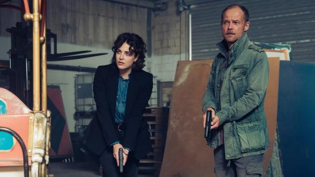 Mann und Frau stehen mit gezogenen Waffen in Lagerhalle vor Gondel eines Riesenrads 