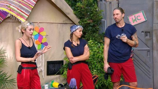 Ariel (l.), Eva Benetatou und Gil Ofarim stehen im Dschungelcamp vor dem WC-Container
