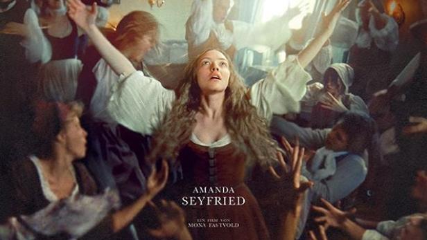 Amanda Seyfried steht mit ausgebreiteten Armen von anderen Personen umgeben da