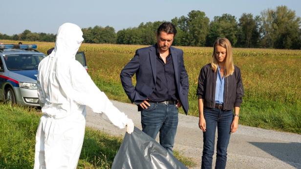 Mann in Schutzanzug hebt Plastikplane und zeigt einem Ermittler-Duo offenbar eine Leiche am Feldrand