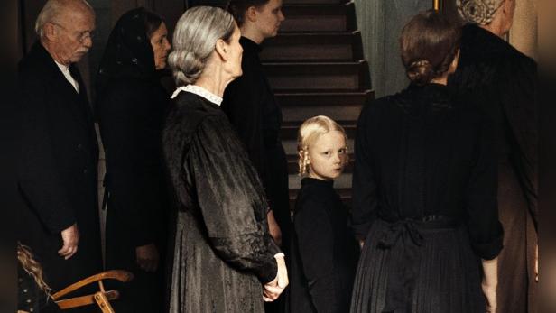 Kleines Mächen in schwarzem Kleid steht inmitten einer Menschengruppe und blickt in die Kamera