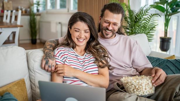 Paar sitzt mit Popcorn vor dem Laptop auf dem Sofa