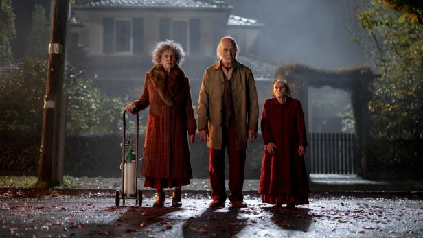 Zwei alte Frauen und ein alter Mann stehen nachts vor einem dunklen Haus auf einer laubbedeckten Straße 
