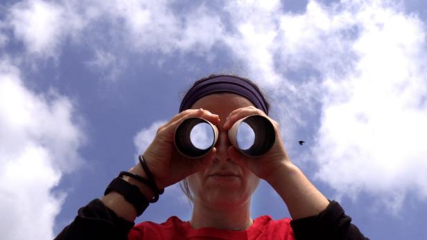Figur mit Fernglas vor Augen, in dem sich der Himmel spiegelt