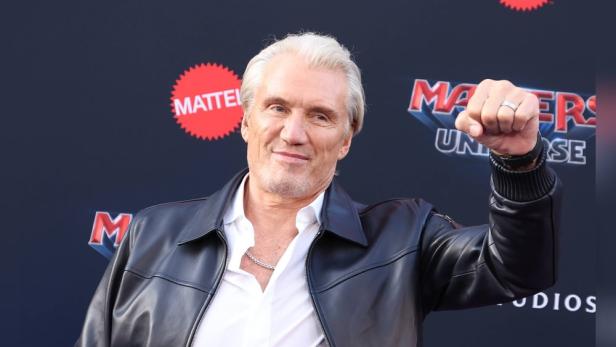 Dolph Lundgren reckt die Faust in die Luft vor einem "Masters of the Universe"-Logo.