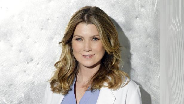 Ellen Pompeo mit blonden, gewellten Haaren und blauem Oberteil unter einem weißen Arztkittel.